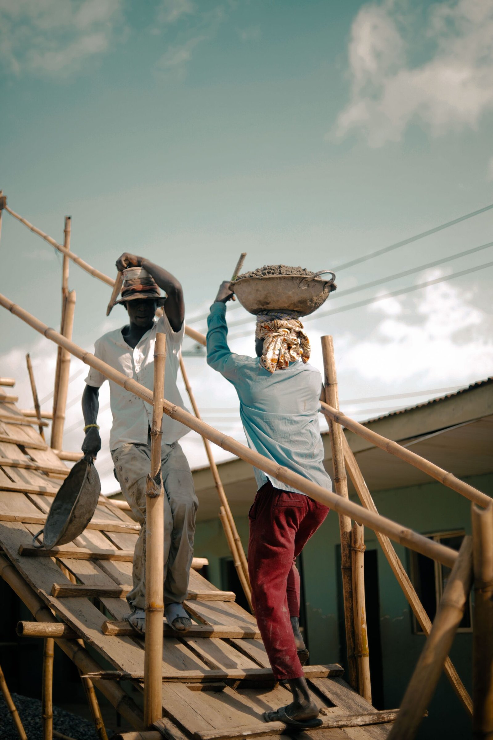 Two African construction workers on a bamboo ramp carrying materials.